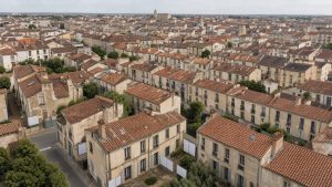 Vue aérienne réaliste d’une ville française moyenne avec maisons anciennes, panneaux immobiliers vierges et centre-ville visible en arrière-plan.